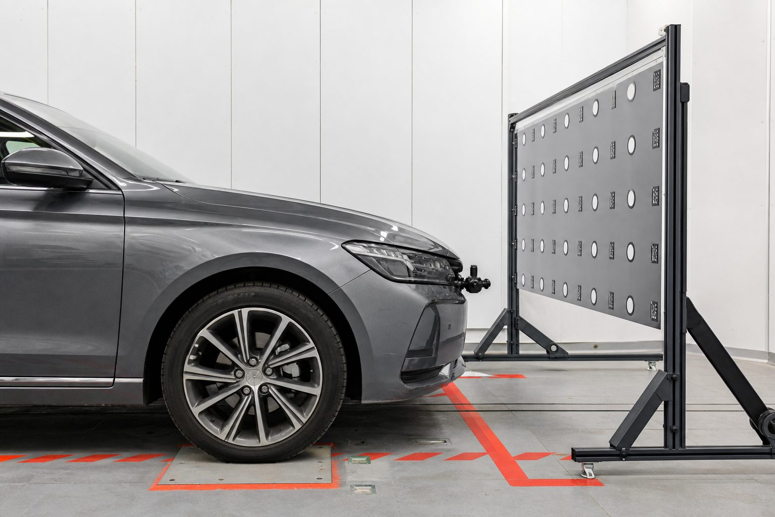 Sedan parked in front of a freestanding calibration target board in a clean white service bay with red floor markings